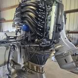 Outboard on a boat transom with its cowling off, intake runners and wiring exposed, a white outboard beside it in a metal-walled shop