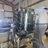 Two outboards on the back of a boat in a metal shop, the left one uncowled with a hoist chain hanging in front of it